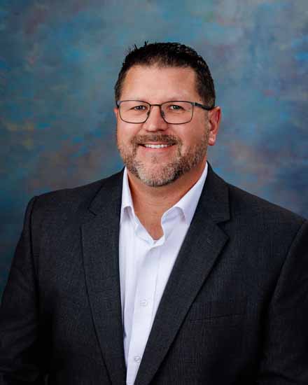 Tim Brown, smiling man in a suit and glasses against a blue background.